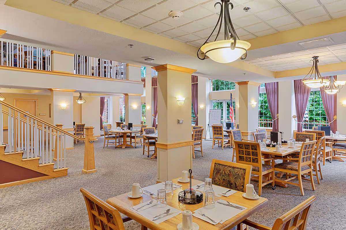 A spacious dining room in an assisted living facility with multiple wooden tables and chairs set with white cups, glasses, and silverware. The room features large windows with purple curtains, beige walls with wooden trim, and hanging light fixtures. There is a staircase leading to an upper level with a railing, and an American flag is visible near the windows.