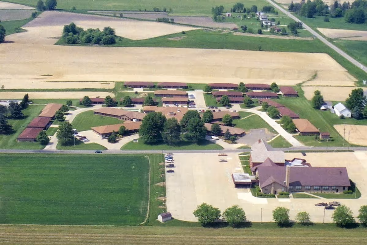 Aerial view of the Apostolic Christian Home facility showing multiple low-rise buildings, a parking lot, and surrounding farmland.