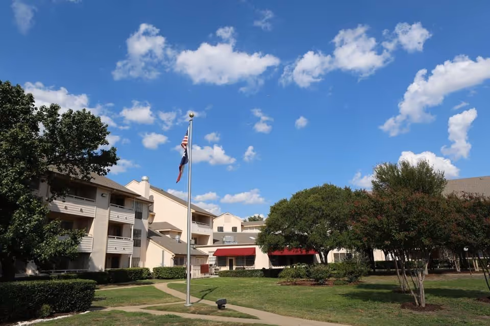 Exterior view of Serenity at Dallas senior living facility showing a multi-story building with balconies, a flagpole with American and Texas flags, green lawn, trees, and a clear blue sky with scattered clouds.
