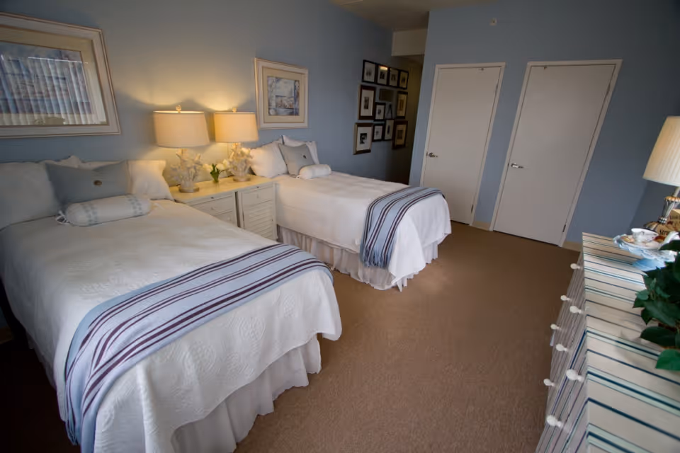 A neatly arranged bedroom with two twin beds, each covered with white bedspreads and striped throws at the foot. Between the beds is a nightstand with two lamps and decorative items. The walls are painted light blue and adorned with framed pictures. There is a dresser with a striped cover and a plant on top, and two closed white doors are visible on the far wall.
