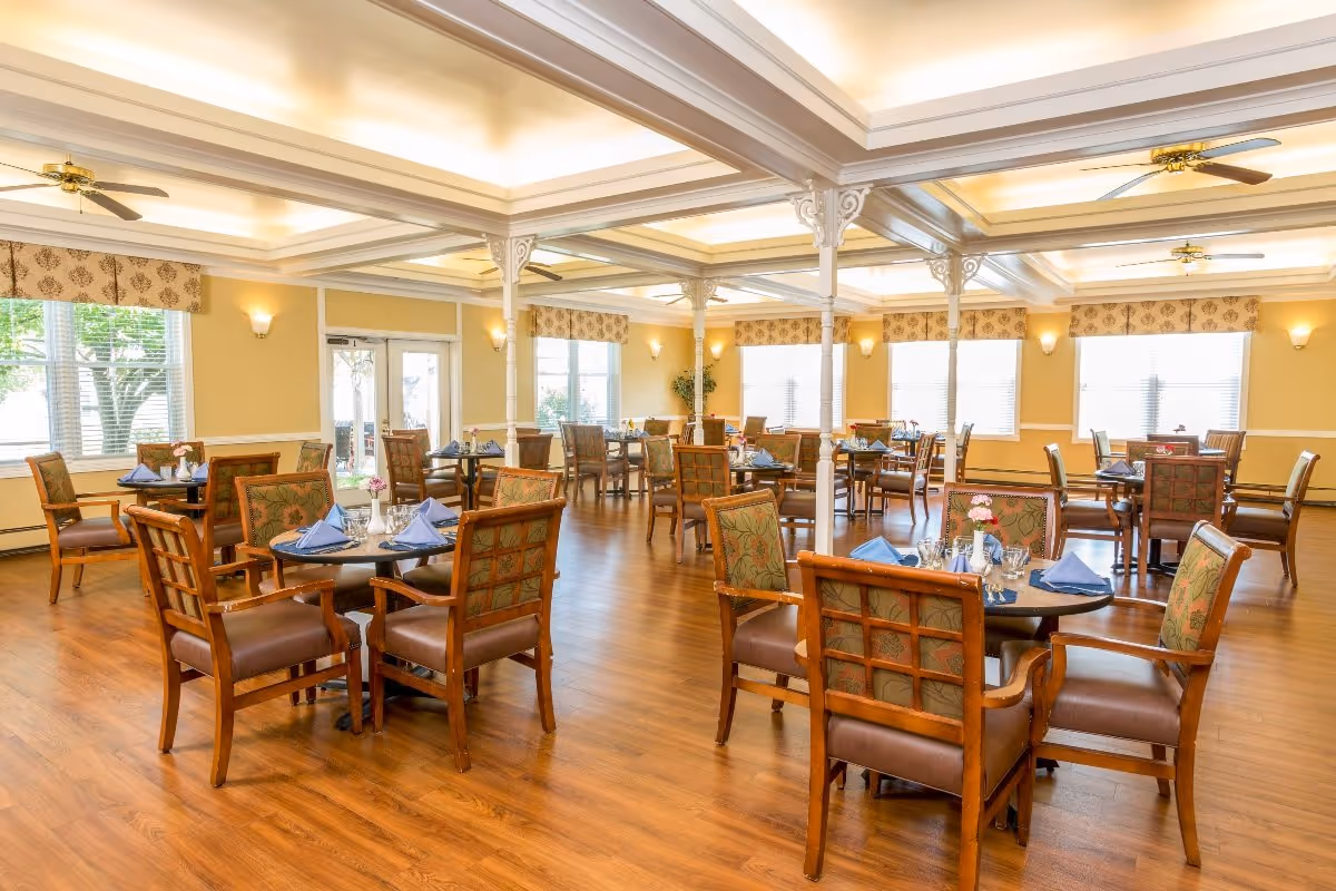 A spacious dining room with multiple round tables set with blue napkins, glassware, and small flower vases. The room has wooden floors, large windows with patterned valances, ceiling fans, and decorative white columns supporting a coffered ceiling.