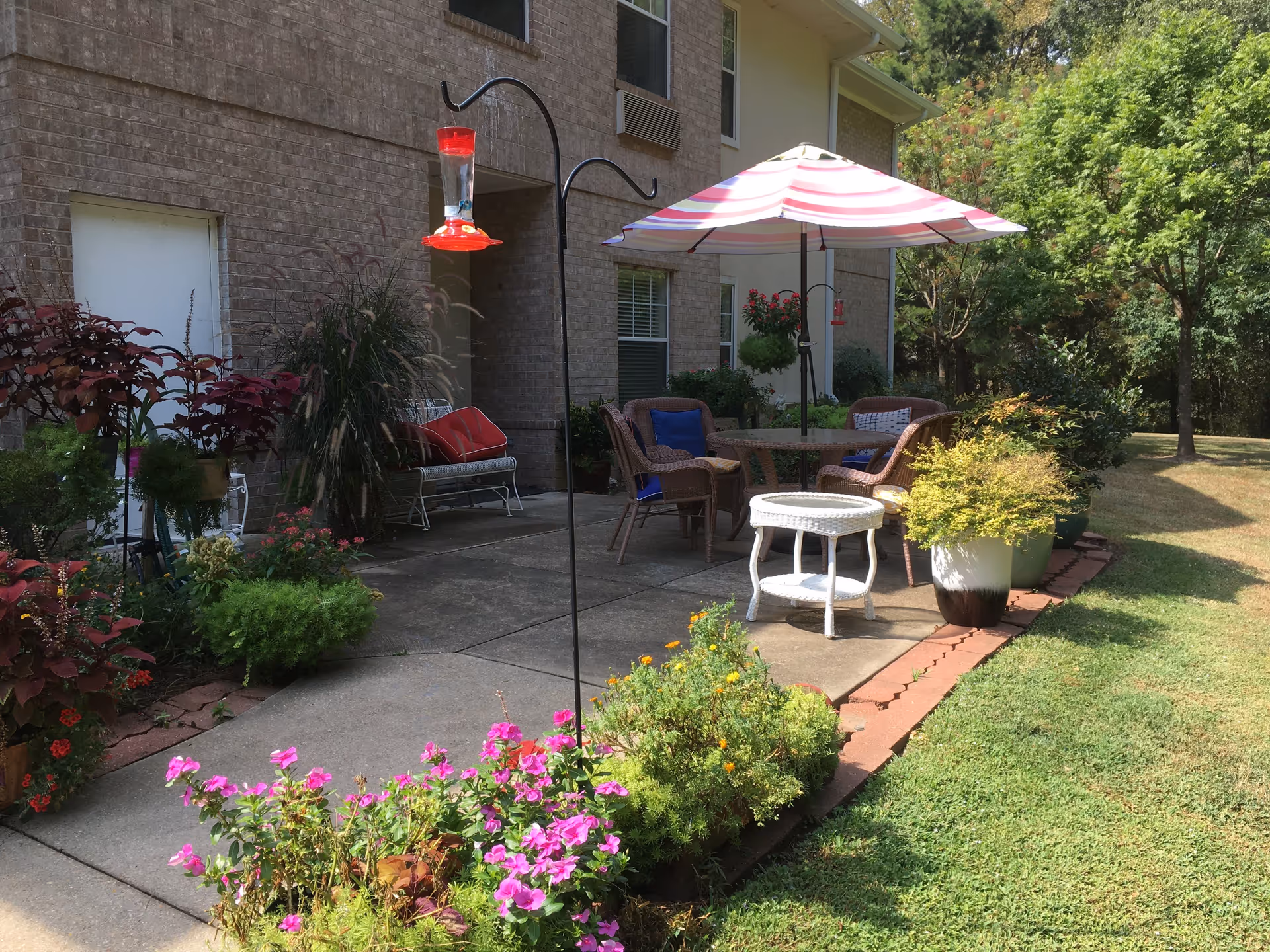 Outdoor patio area with a round table and four wicker chairs, some with blue cushions, under a pink and white striped umbrella. The patio is surrounded by various potted plants and flowers, with a brick building wall in the background and a green lawn and trees to the right.
