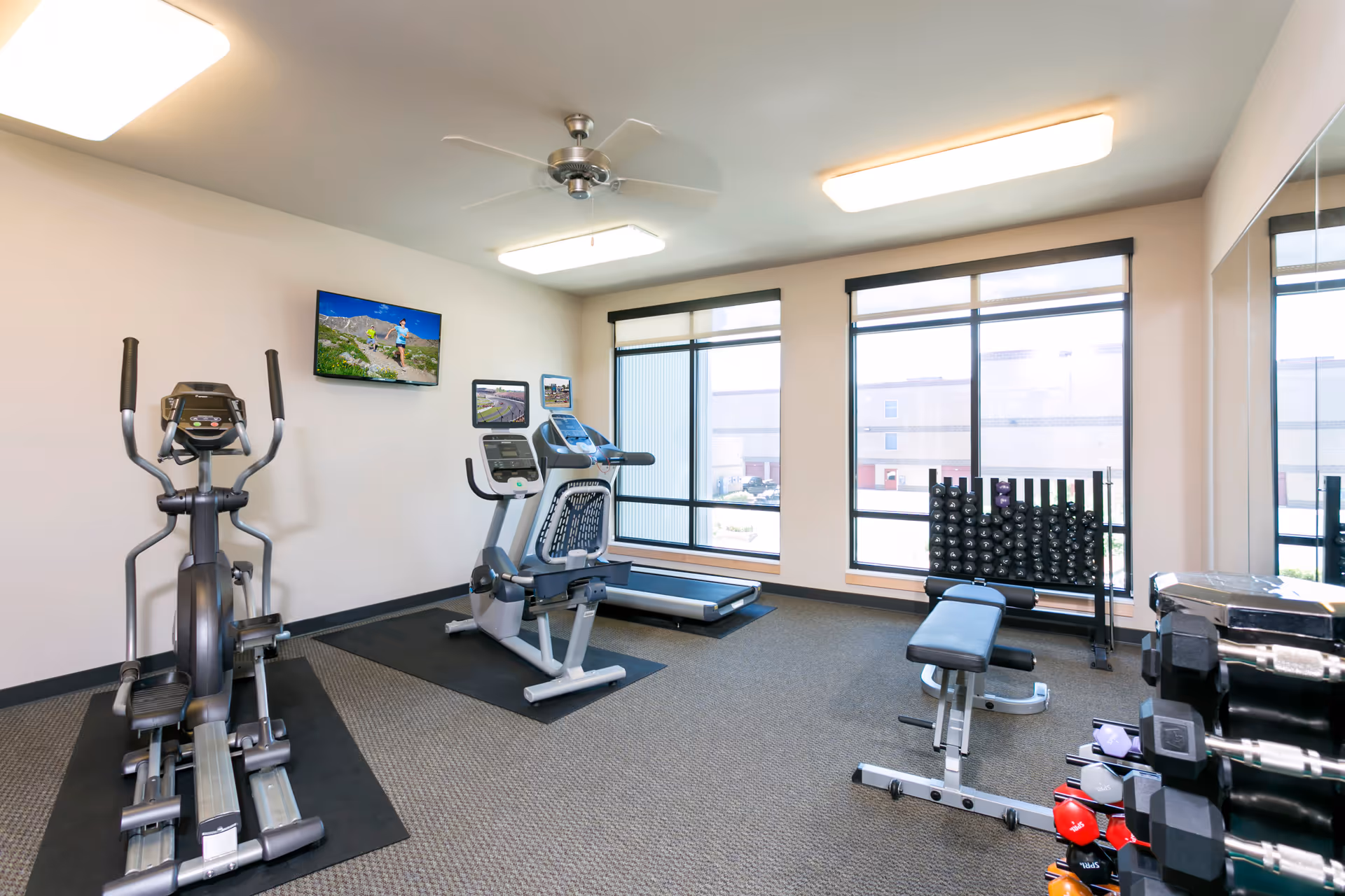 A bright fitness room with exercise equipment including an elliptical machine, a recumbent bike, a treadmill, a weight bench, and a rack of dumbbells. Large windows let in natural light, and two flat-screen TVs are mounted on the wall. The room has a ceiling fan and overhead lighting.