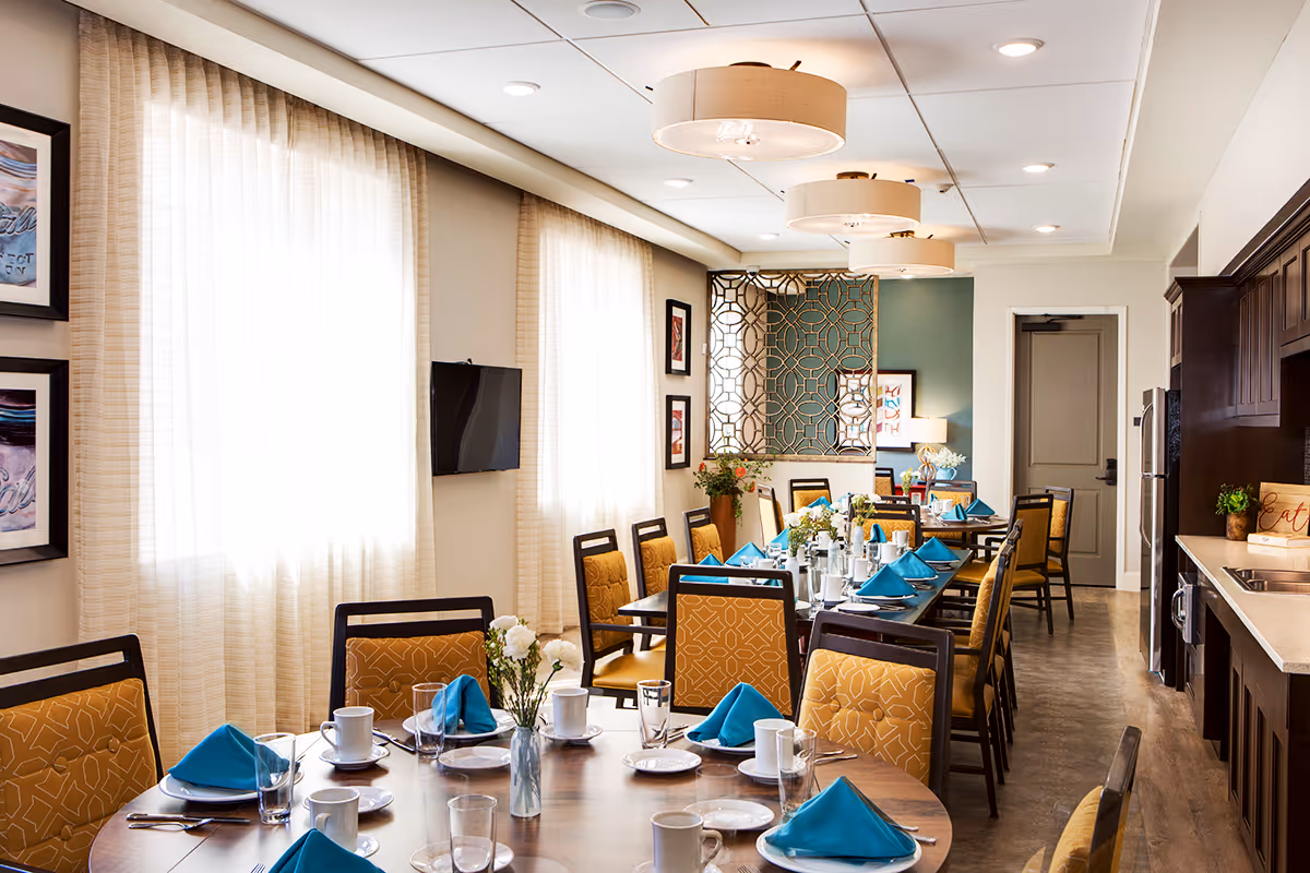 A bright and elegant dining room in The Legacy at South Plains facility, featuring multiple tables set with white plates, cups, glasses, and blue folded napkins. The room has large windows with sheer curtains, yellow cushioned chairs, decorative wall art, and modern ceiling lights. A kitchenette area with dark wood cabinets and a countertop is visible on the right side.