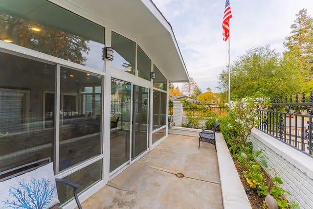 Outdoor patio area with a concrete floor, black metal bench, and a chair with a blue coral-patterned cushion. The patio is adjacent to a building with large glass windows and doors. There is a white brick planter with green plants and a black metal fence. An American flag is visible on a flagpole in the background under a partly cloudy sky.