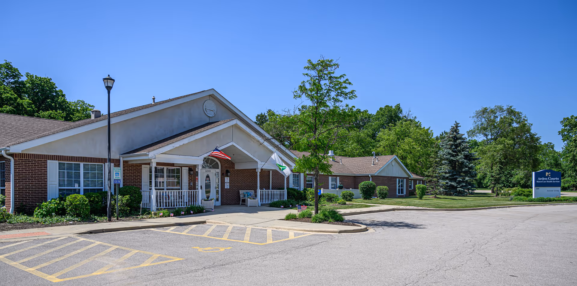 Exterior view of Arden Courts - ProMedica Memory Care Community in Northbrook, showing a single-story brick building with a covered entrance, an American flag, a small tree, and a parking area with marked spaces. There is a blue sign on the lawn indicating the facility name.