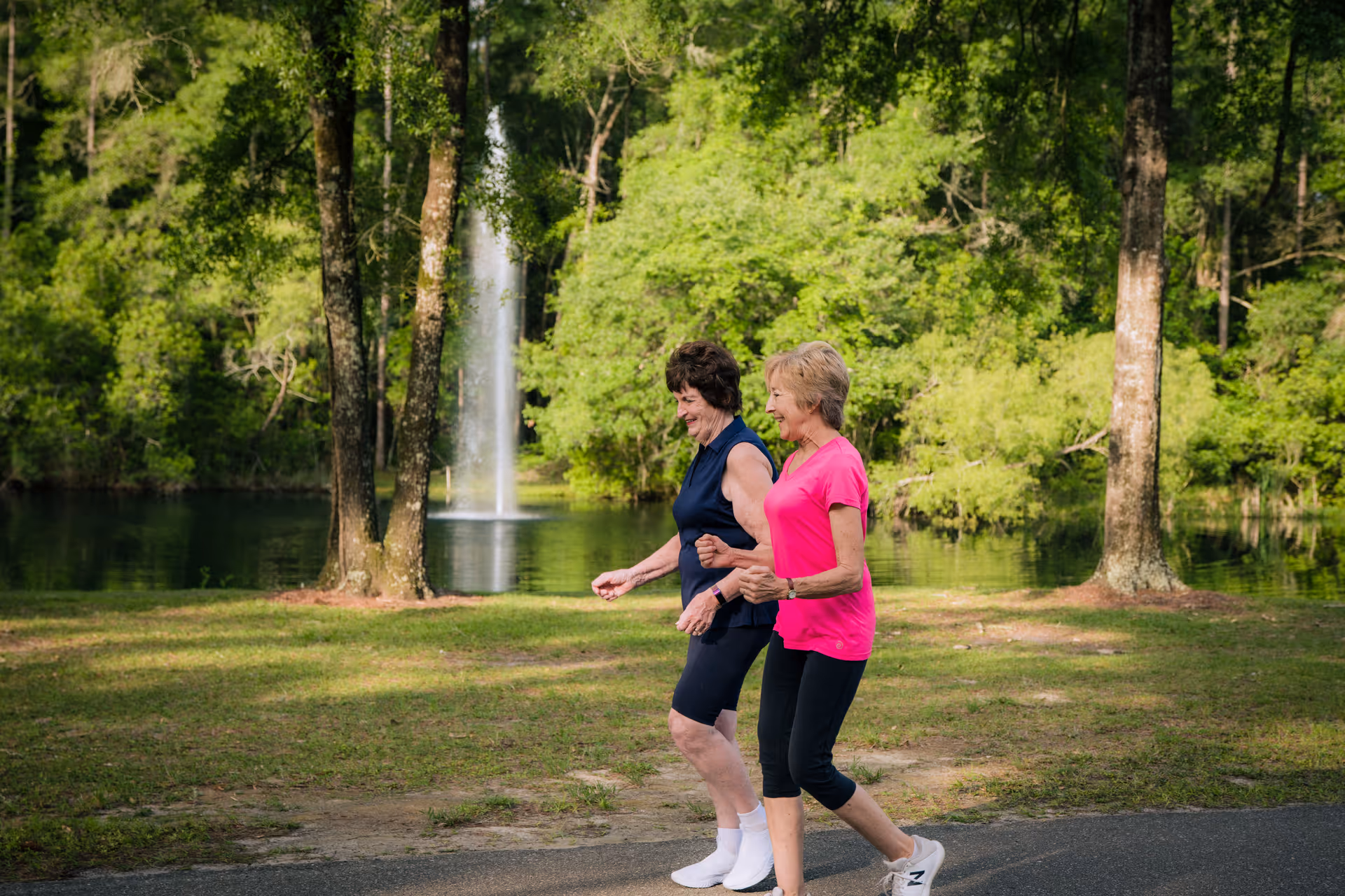 Two elderly women walking and smiling together on a paved path near a pond with a water fountain, surrounded by green trees and grass in an outdoor park-like setting.