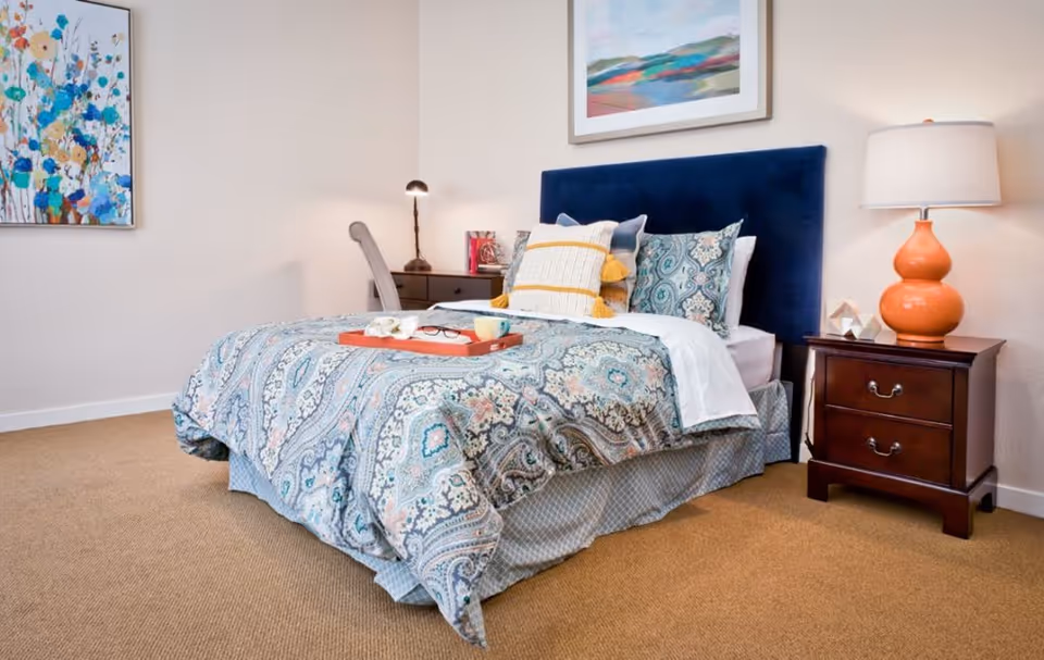 A neatly staged bedroom with a patterned bedspread, navy upholstered headboard, bedside tables and lamps.