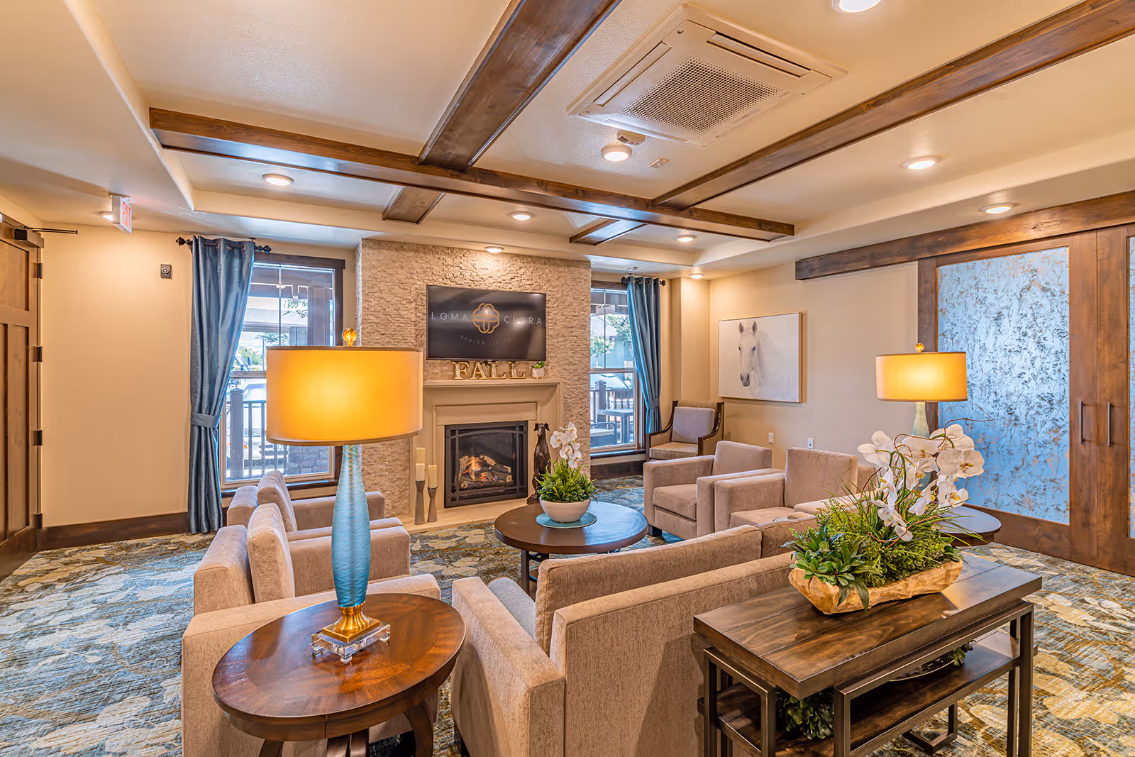 Cozy senior living lounge with upholstered chairs arranged around a fireplace, lamps, and decorative plants.
