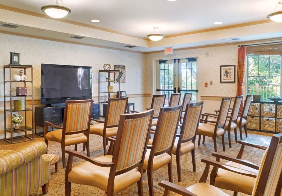 Communal activity room with rows of striped upholstered chairs facing a large flat-screen TV and glass doors to the outside.