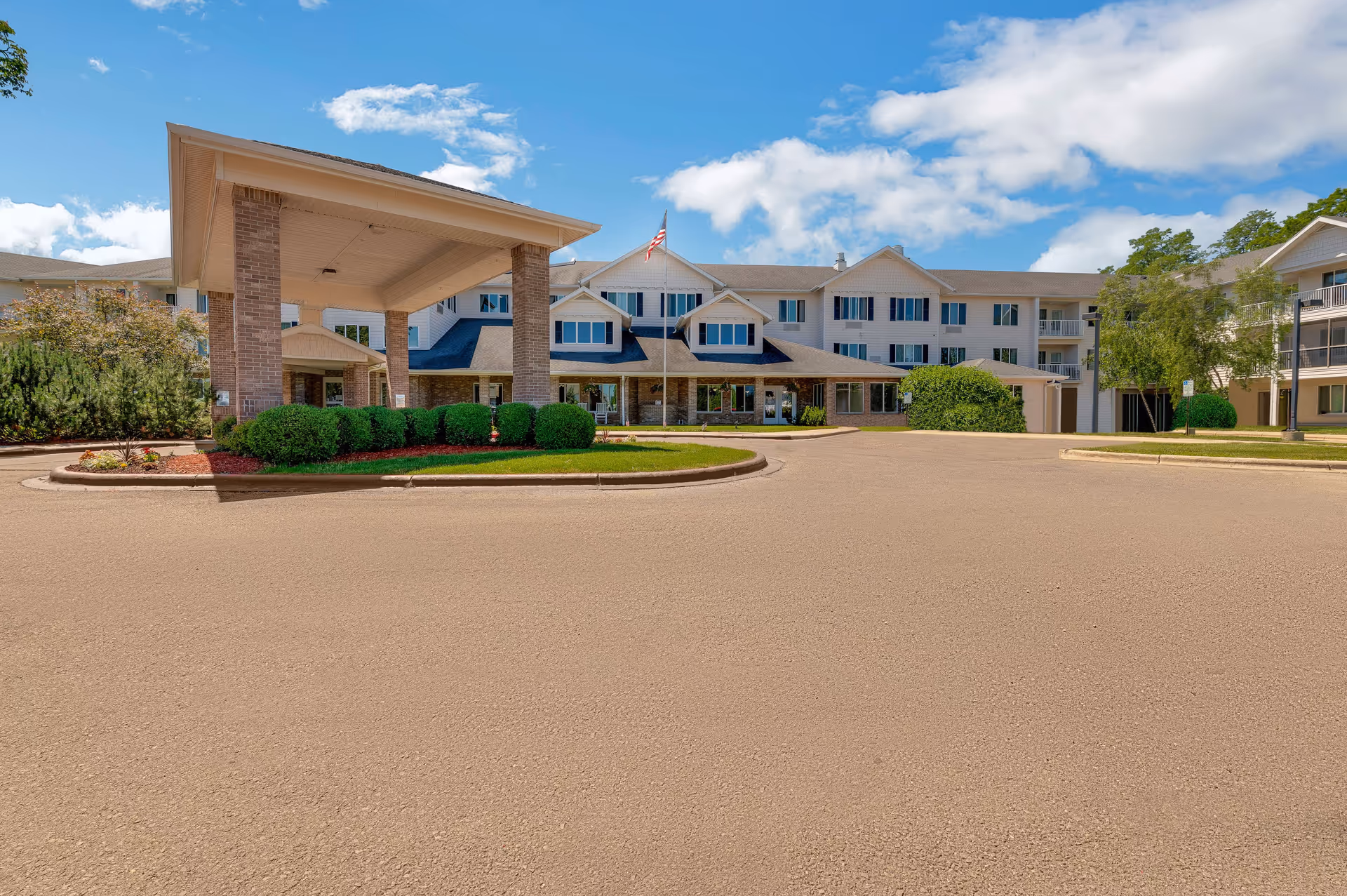 Front exterior of a multi-story senior living building with a covered porte-cochere, circular driveway, landscaping and an American flag.