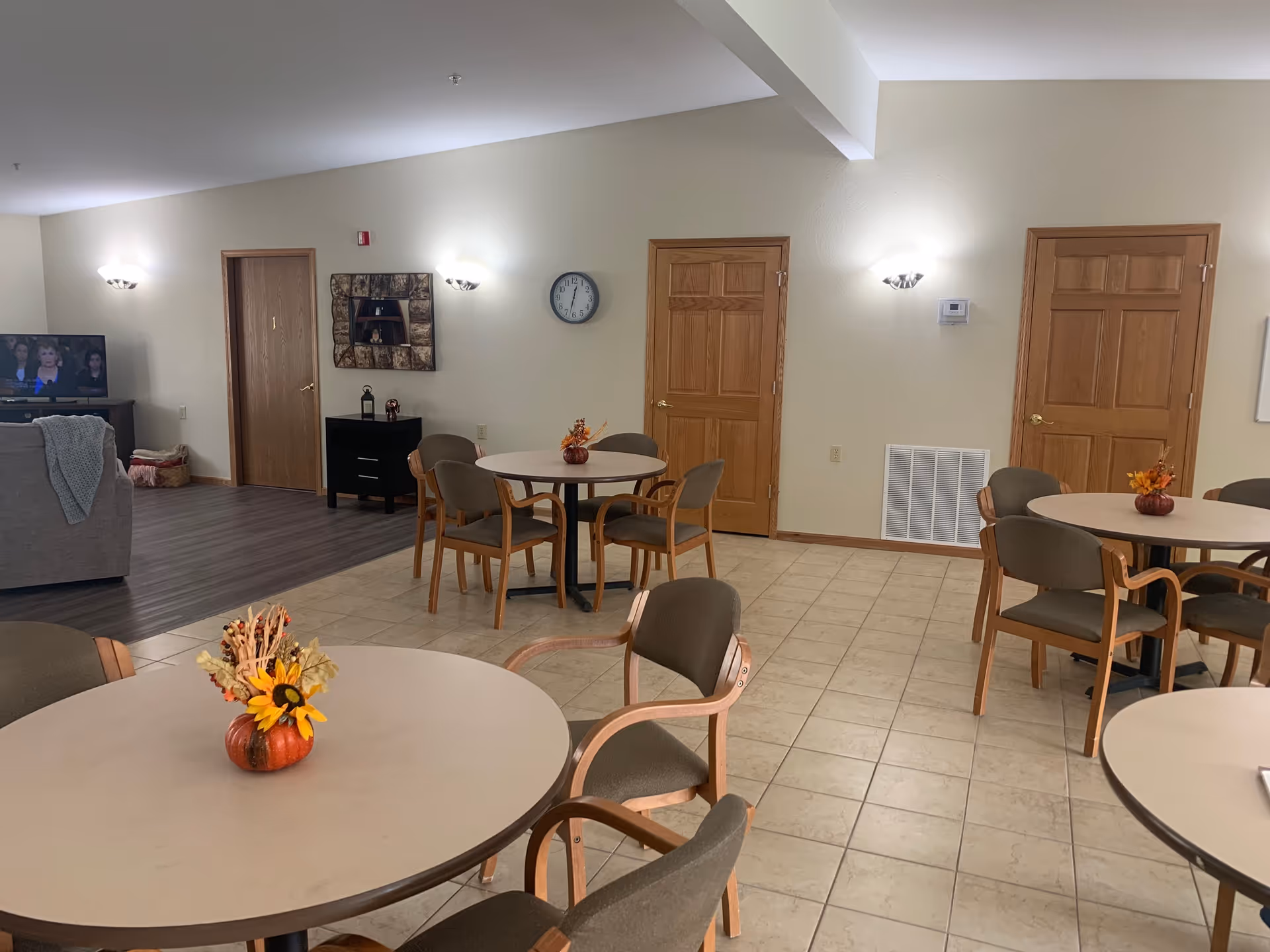 A communal dining area with round tables and chairs arranged on a tiled floor. Each table has a small autumn-themed centerpiece with flowers and leaves. The room has beige walls with wooden doors and wall-mounted lights. In the background, there is a living area with a television and a couch.