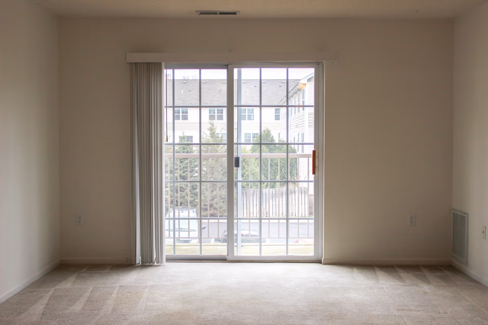 Empty carpeted living room with sliding glass patio doors and vertical blinds looking out to an apartment courtyard.
