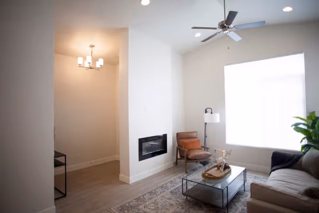 A bright living room with a large window covered by a white blind, a ceiling fan with lights, a modern wall-mounted electric fireplace, a brown leather armchair, a floor lamp, a glass coffee table with decorative items, a patterned area rug, and a beige sofa with a dark throw blanket. The room has light-colored walls and wood flooring.