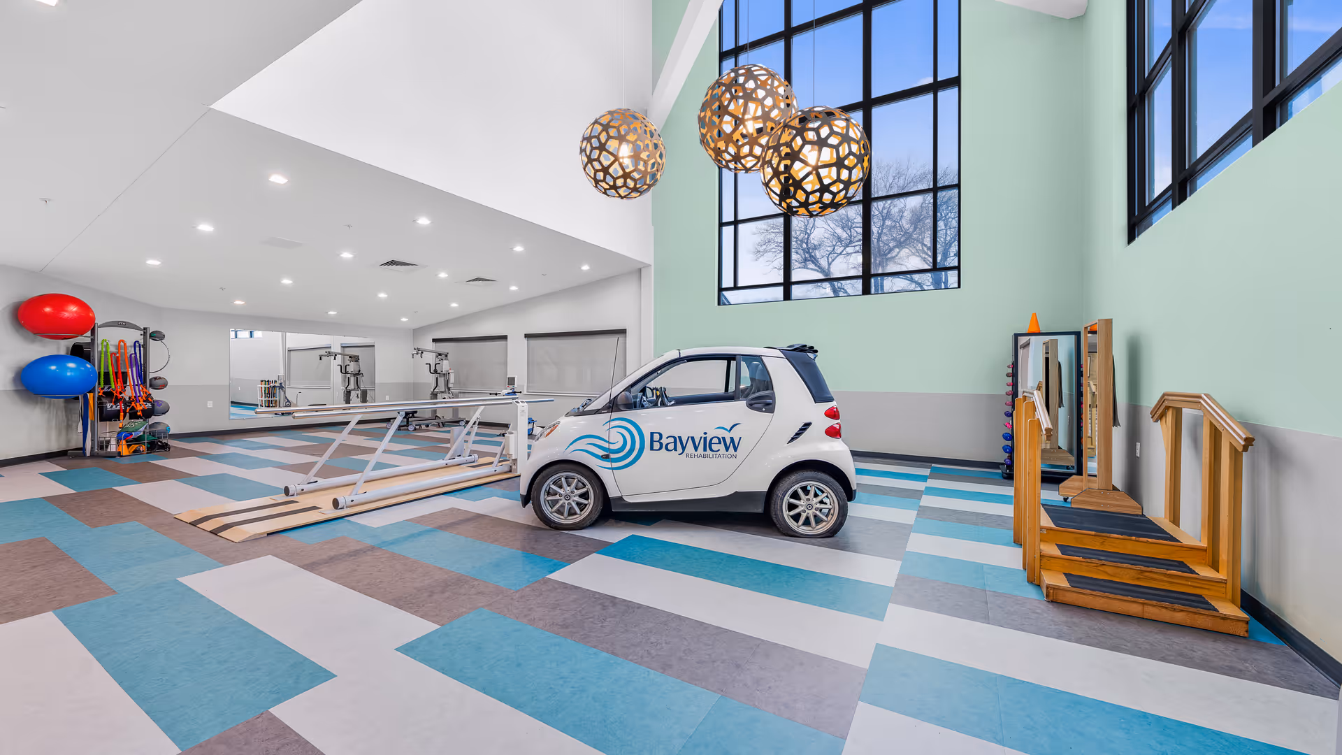 A spacious rehabilitation room with colorful patterned flooring, large windows letting in natural light, and modern pendant lights. The room contains a small white car with Bayview Rehabilitation branding, exercise equipment including balance balls, resistance bands, parallel bars for walking practice, and wooden stairs with handrails for physical therapy.