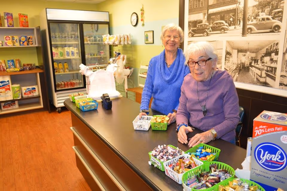 Two elderly women smiling and standing behind a counter with baskets of assorted candy and snacks in a small store or snack area. Shelves with various packaged goods and a refrigerated display case are visible in the background.