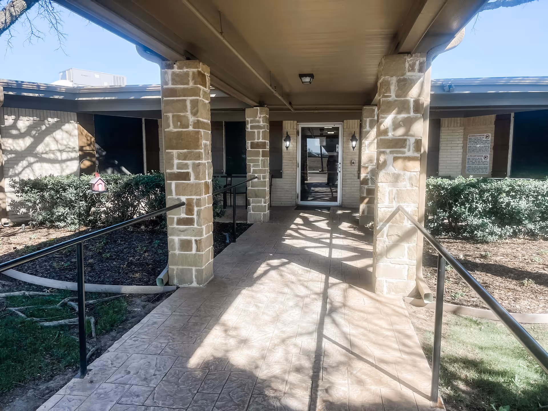 Covered entrance walkway with stone pillars and handrails leading to glass doors, flanked by shrubs.