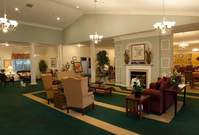 A spacious and warmly lit senior living facility common area with green carpet and beige accents. The room features comfortable seating including armchairs and sofas arranged around wooden coffee tables. There is a decorative fireplace with a 'WELCOME' sign above it, framed artwork, and floral arrangements. Chandeliers hang from the ceiling, and there are columns and plants adding to the inviting atmosphere.