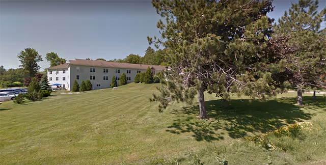 White three-story building set behind a wide mowed lawn with scattered trees under a clear sky.