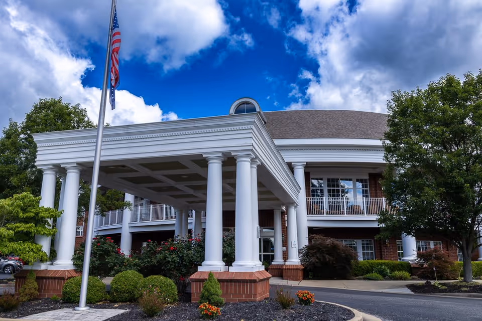 Front exterior view of Elison Independent & Assisted Living of Maplewood building with white columns supporting a covered entrance, an American flag on a flagpole, well-maintained landscaping with bushes and flowers, and a partly cloudy blue sky.