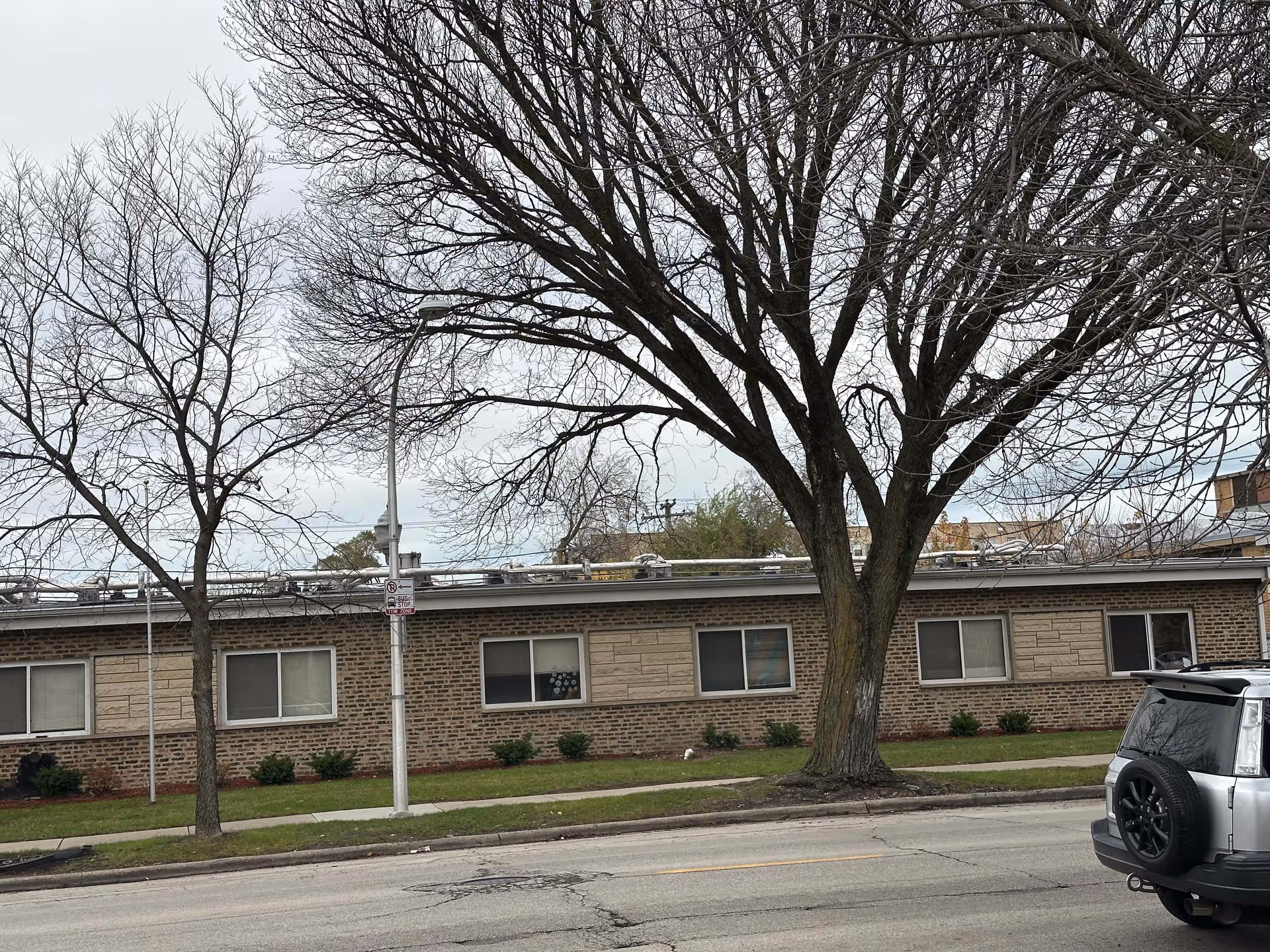 One-story brick nursing home building facade with large leafless trees and a parked SUV along the street.