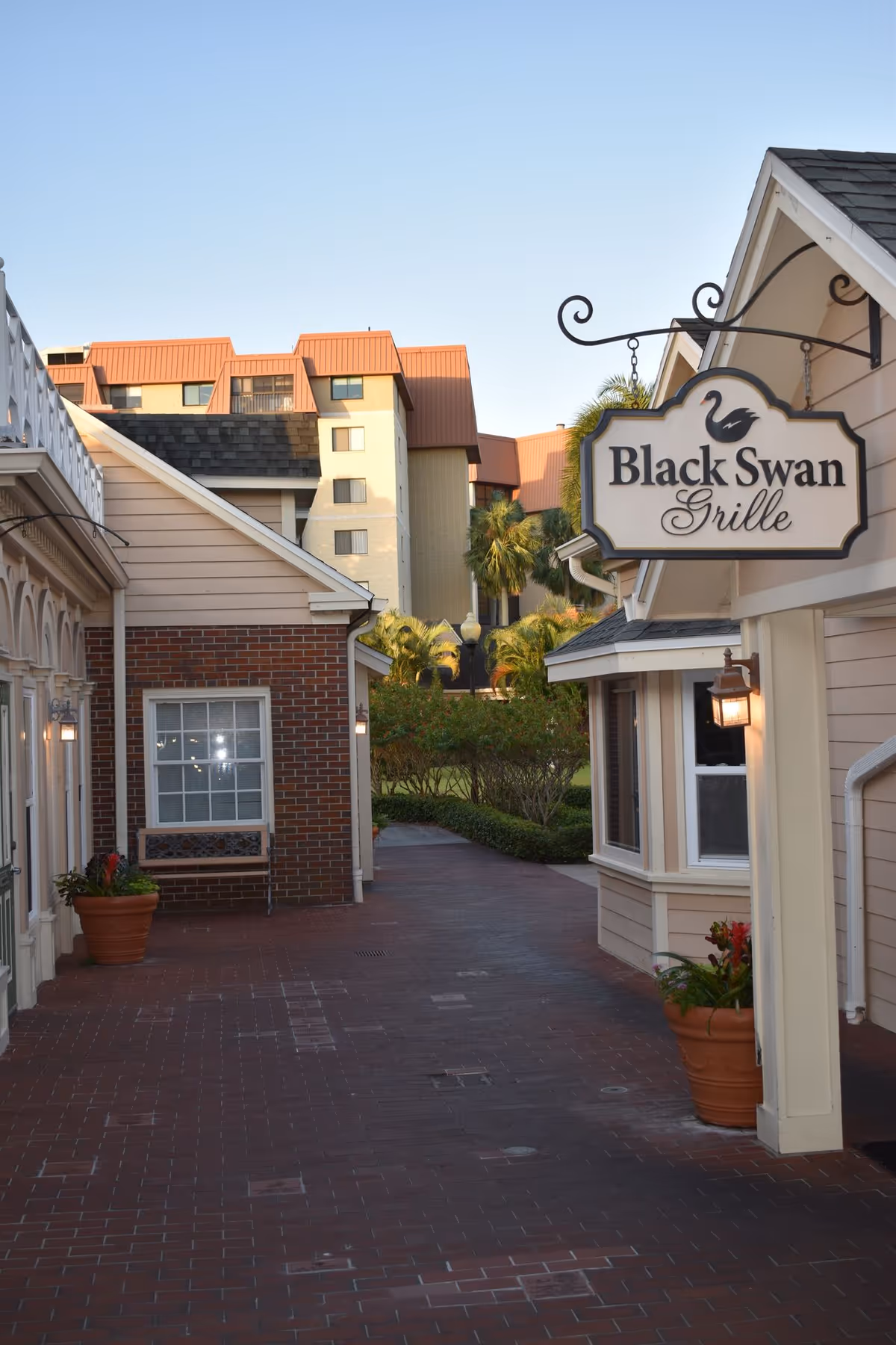 Outdoor walkway between buildings with brick and siding exteriors, potted plants, and a hanging sign for Black Swan Grille restaurant, with taller buildings and palm trees in the background under a clear sky.