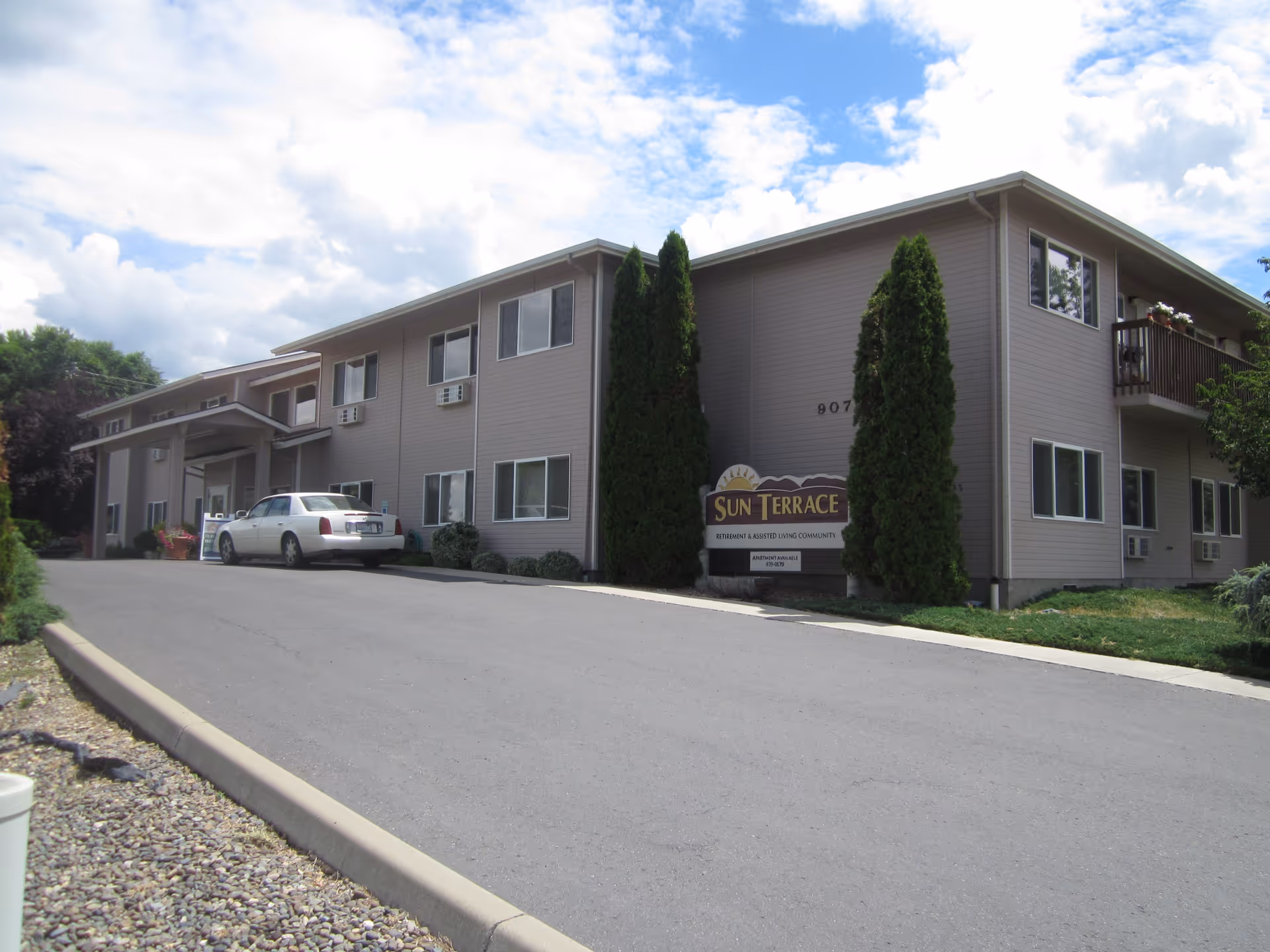 Two-story beige assisted living building front with a driveway, parked car, landscaping and a Sun Terrace sign.