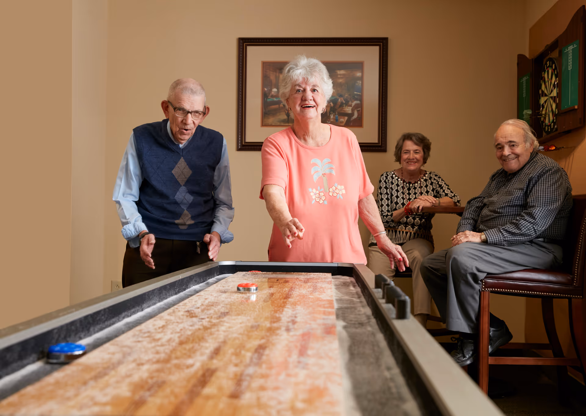 Four elderly individuals enjoying a game of shuffleboard indoors. Two are actively playing while the other two sit nearby watching and smiling. The room has beige walls, a framed picture, and a dartboard on the wall.