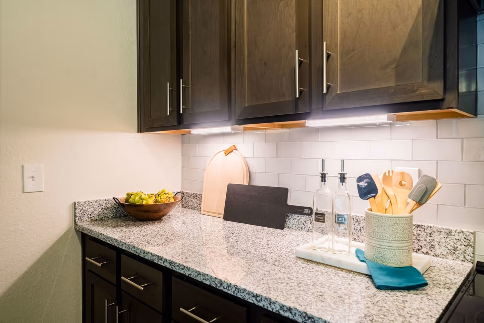 Granite kitchen countertop with dark cabinets, cutting boards, a bowl of fruit, oil bottles and utensils in a crock.