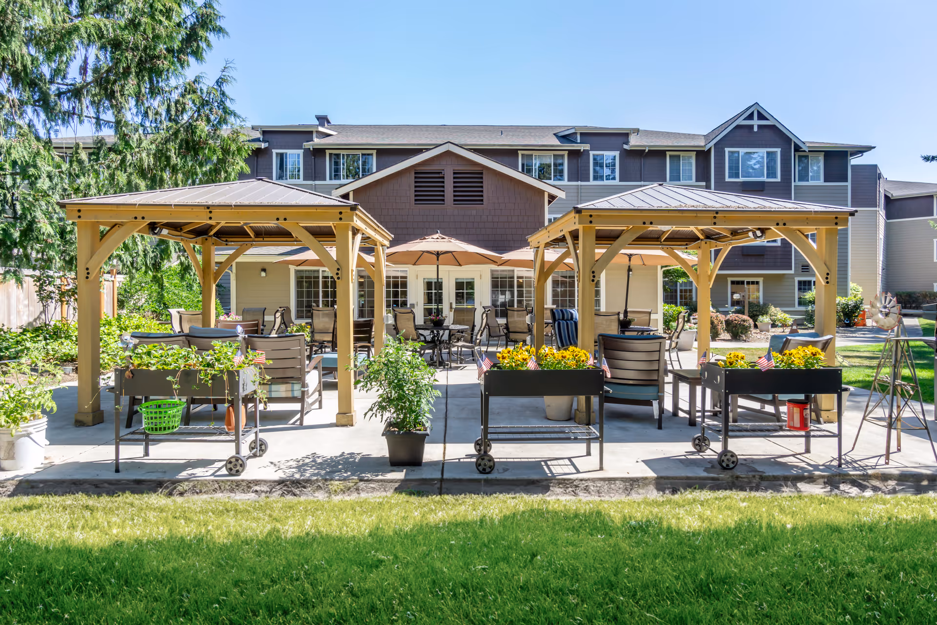 Outdoor patio area at Brookdale Courtyard Puyallup featuring two wooden gazebos with seating underneath, several potted plants and flowers, patio tables with umbrellas, and a multi-story residential building in the background under a clear blue sky.