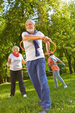 Three elderly people outdoors in a grassy area with trees, smiling and playing with hula hoops on a sunny day.