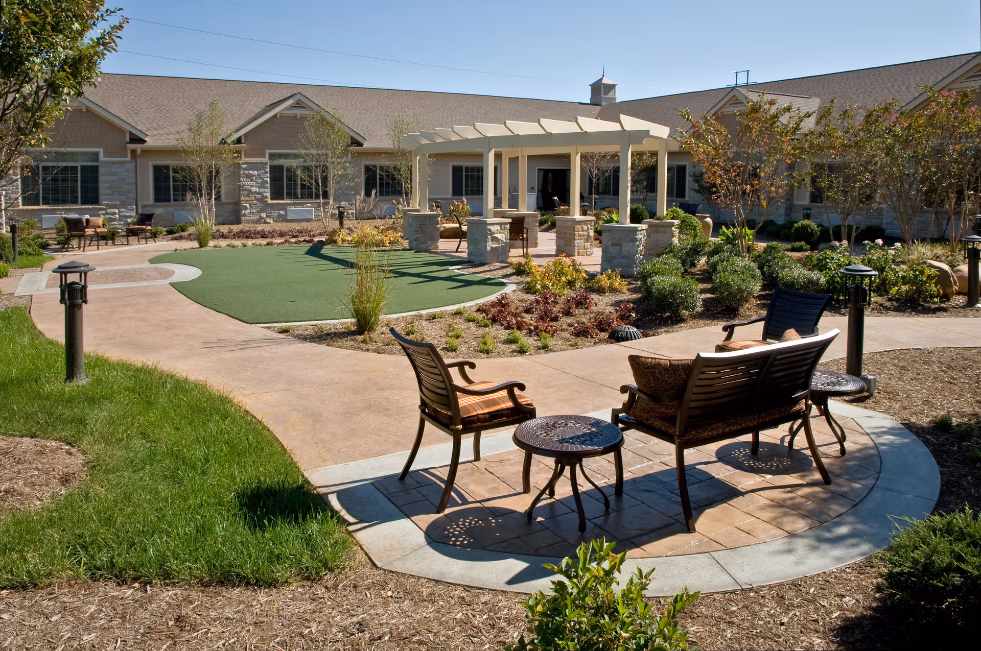 Outdoor courtyard area at a senior living facility with paved walkways, green lawn, shrubs, trees, and seating including chairs and a bench under a pergola structure in front of a single-story building.