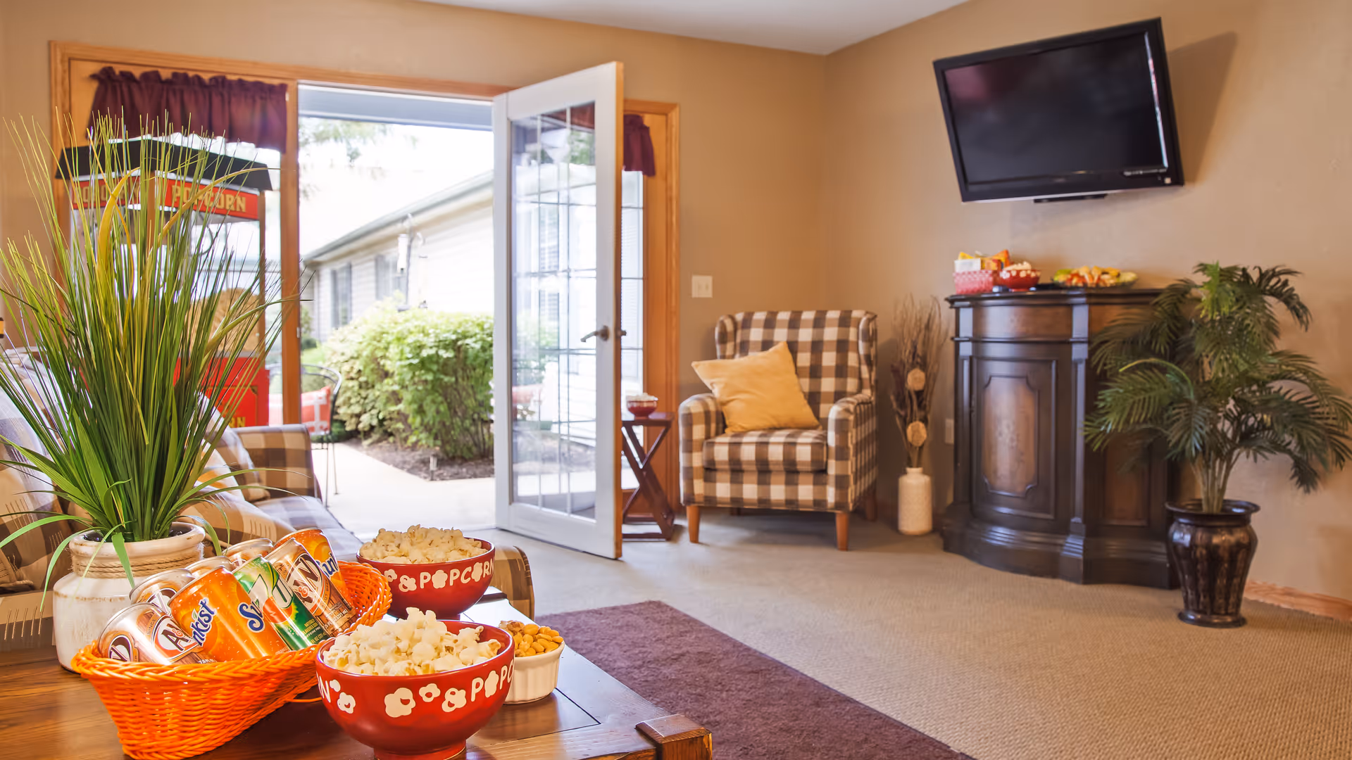 A cozy living room area with a checkered armchair, a small wooden table, a wall-mounted TV, and a cabinet with snacks on top. There is a basket with soda cans and bowls of popcorn and nuts on a wooden coffee table in the foreground. The room has beige walls and carpet, with a glass door open to an outdoor area with greenery.