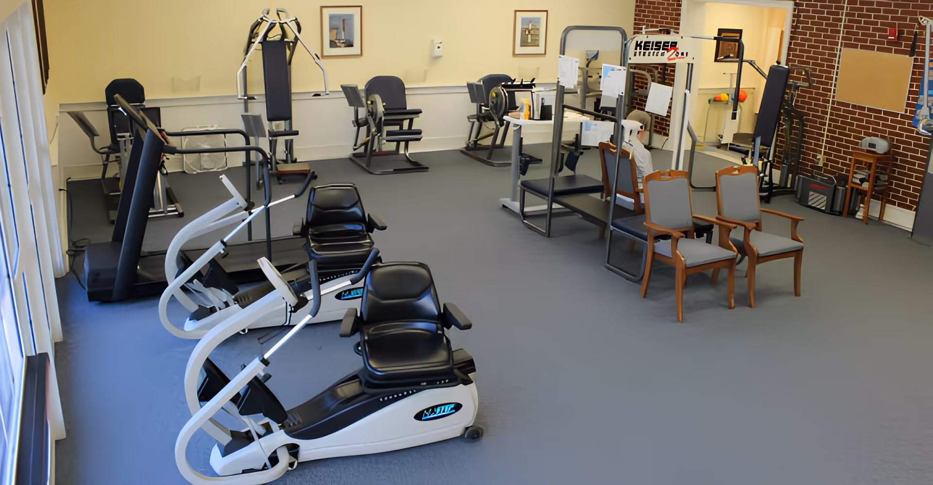 Exercise/fitness room in a retirement community with recumbent bikes, strength machines, and a few chairs.