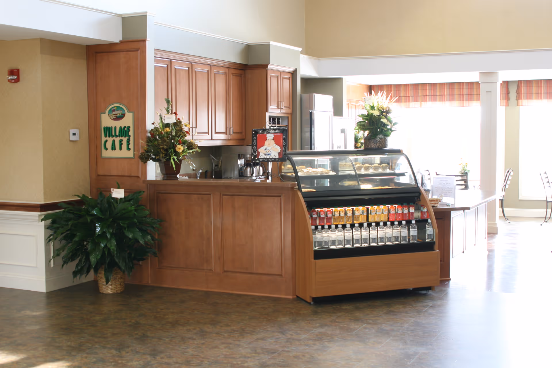 Interior view of a café area named Village Café with wooden cabinetry, a display case filled with beverages and pastries, floral arrangements on the counter, and tables and chairs in the background near large windows with striped valances.