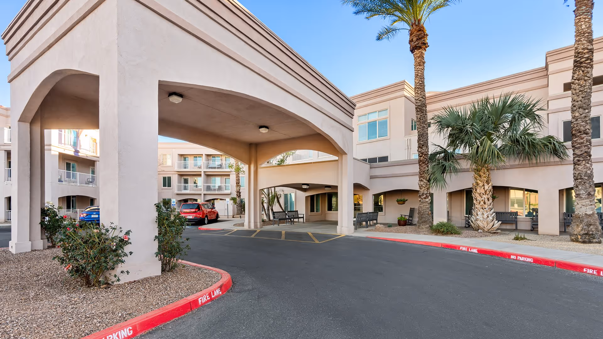 Exterior view of Holiday Vista de la Montana senior living facility entrance with covered drop-off area, palm trees, benches, and parked cars under a clear blue sky.
