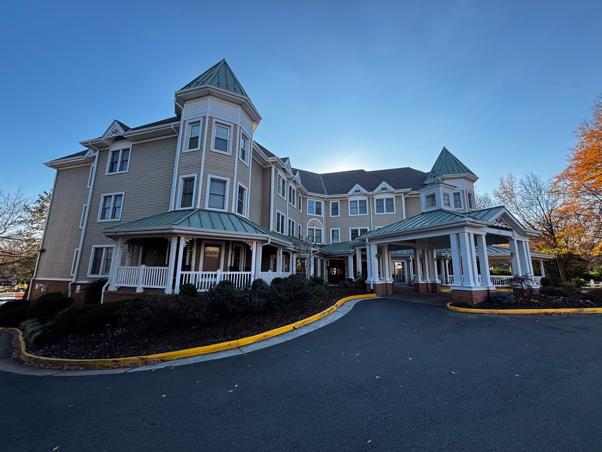 Front exterior of a multi-story senior living building with a covered porte-cochere and decorative turrets under a clear blue sky.