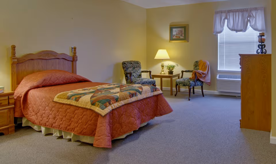 A cozy bedroom with a wooden bed frame and a quilted rust-colored bedspread. There is a patchwork quilt folded at the foot of the bed. Two floral upholstered chairs flank a small wooden side table with a lamp and a small plant. A framed picture hangs on the wall above the table. A window with blinds and a valance is next to a wooden dresser with decorative items on top. The room has beige walls and carpeted flooring.