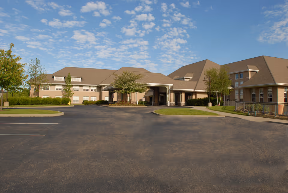Exterior view of a large senior living facility building with a covered entrance, surrounded by trees and landscaping under a partly cloudy blue sky.