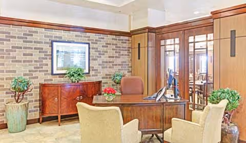 A reception-style interior with a wooden desk, upholstered chairs, brick accent wall, framed art and potted plants.