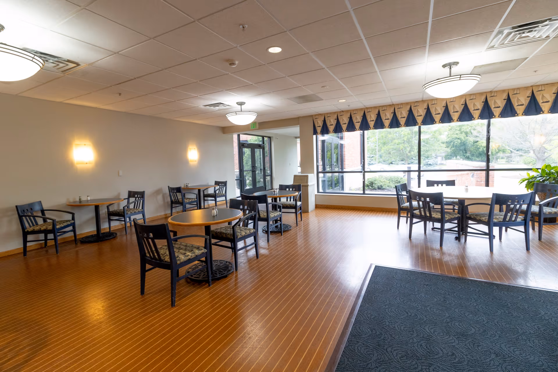 A bright dining area with several round tables and chairs arranged on a wooden floor. Large windows on one side let in natural light and offer a view of greenery outside. The ceiling has recessed lighting and two hanging light fixtures. The walls are light-colored with two wall sconces providing additional lighting. There is a dark entrance mat near the door.