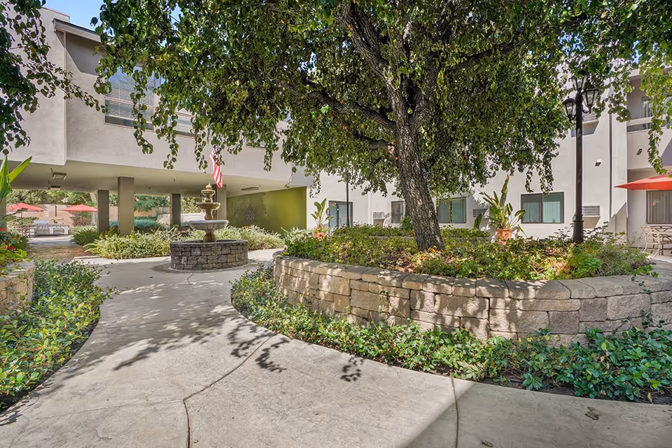 Shaded courtyard featuring a large tree, raised stone planters, a tiered fountain, and walkways surrounded by building facades.