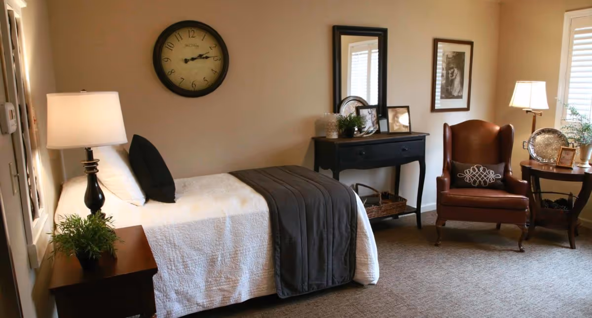 A cozy bedroom with a single bed covered in a white quilt and a dark gray blanket. There is a black pillow and a white pillow on the bed. A wooden nightstand with a lamp and a small plant is beside the bed. On the wall above the bed is a round clock. Across from the bed is a black console table with a mirror, framed pictures, and decorative items. To the right, there is a brown leather armchair with a decorative pillow, a round wooden side table with a lamp, a silver tray, and a small plant. The room has beige walls and carpeted floor with windows letting in natural light.