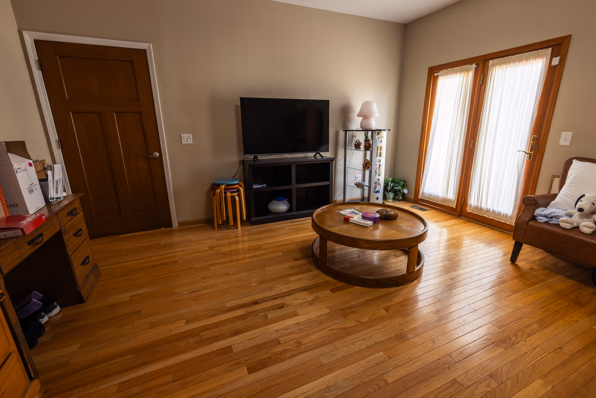 A cozy living room with wooden flooring, a round wooden coffee table in the center, a TV on a black stand against the wall, a glass display cabinet with decorative items, a brown armchair with a white pillow and a stuffed toy, and double glass doors with sheer curtains letting in natural light.