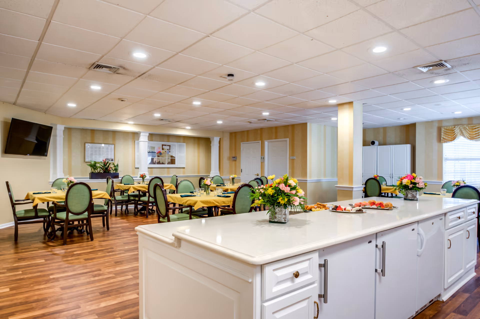 A spacious dining room with multiple tables covered in yellow tablecloths and green cushioned chairs. A large white island counter in the foreground holds flower arrangements and plates of food. The room has wood flooring, beige striped walls, recessed ceiling lights, and a wall-mounted TV.