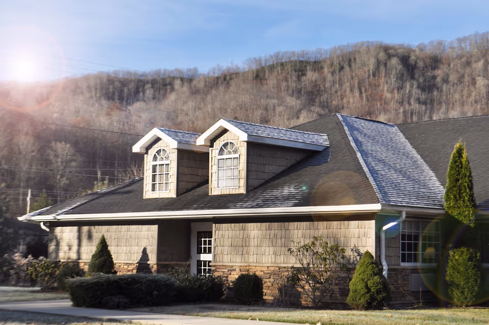 Exterior view of a single-story building with two dormer windows on the roof, surrounded by shrubs and small trees, with a backdrop of wooded hills under a clear sky.