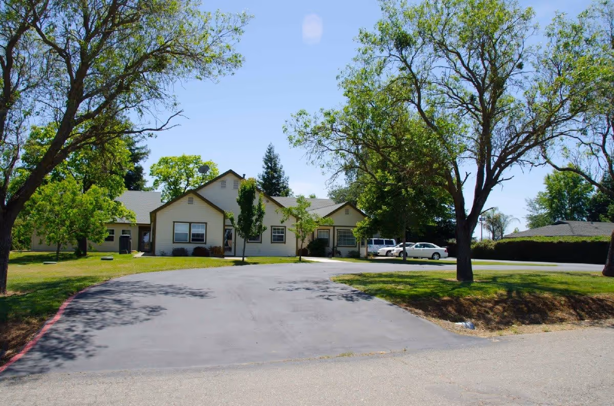 Exterior view of a single-story building with beige siding and multiple windows, surrounded by green trees and a paved driveway leading up to the entrance. Several cars are parked to the right side of the building under a clear blue sky.