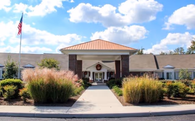 Front exterior view of a single-story building with a covered entrance, landscaped bushes, and an American flag on a flagpole under a partly cloudy sky.