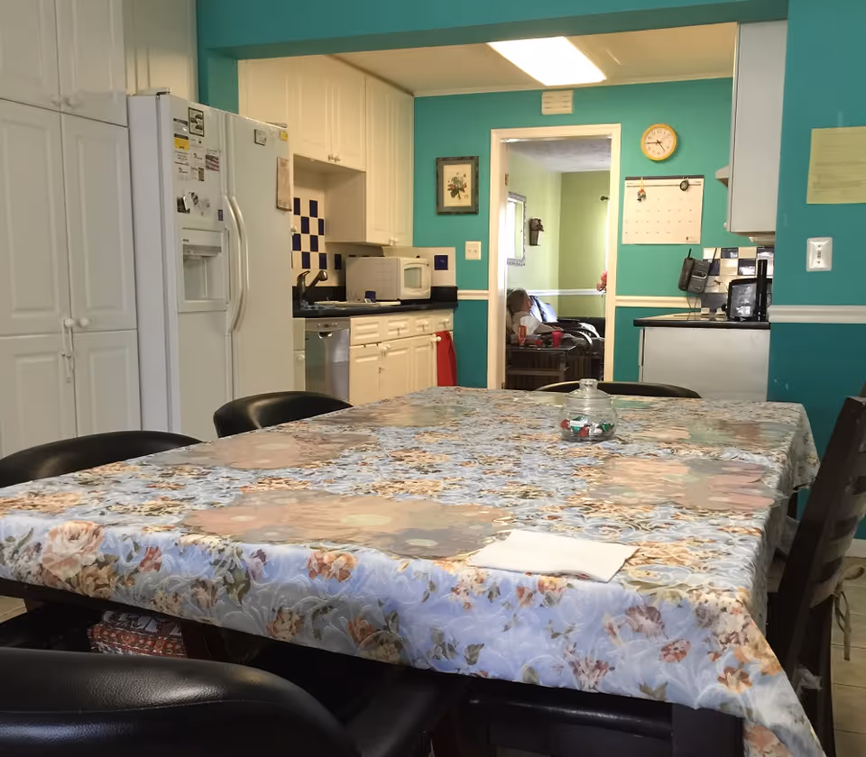 A kitchen and dining area with a large table covered by a floral tablecloth surrounded by black chairs. The kitchen has white cabinets, a white refrigerator, a microwave, and a dishwasher. The walls are painted teal with white trim. In the background, there is a doorway leading to a living room with a person sitting on a couch.