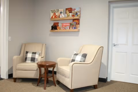 Two beige armchairs with black and white checkered pillows are positioned around a small wooden round table in a room with light-colored walls and carpeted floor. A white door with a silver handle is visible on the right side, and two wooden shelves mounted on the wall above the chairs hold various books and decorative items.